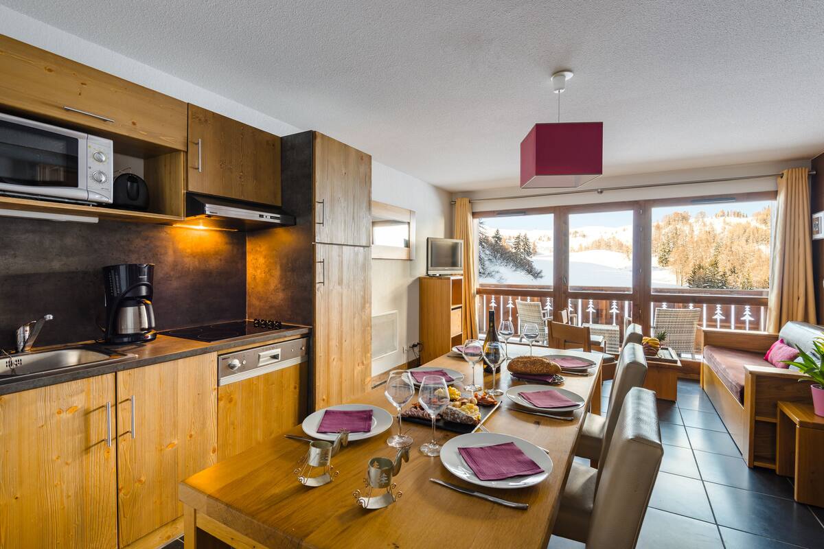 A modern kitchen with wooden cabinets, a dining area with a wooden table set for four, and a balcony with a view of a snowy landscape.