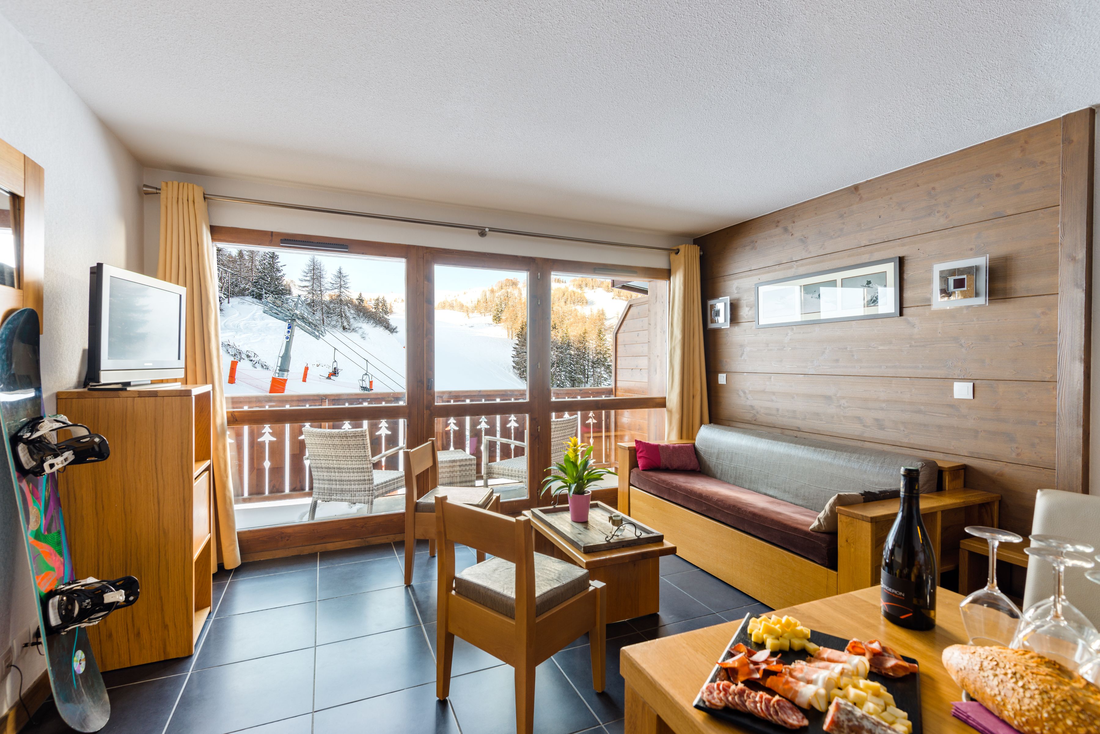 A cozy room with a wooden floor, a sofa, a dining table with a board game, a TV, and a ski slope view.