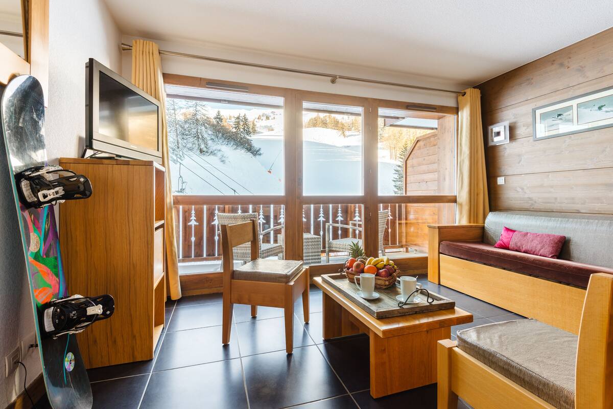 A cozy room with a wooden sofa, a dining table with fruit and a cup, a TV on a stand, and a snowboard leaning against the wall.