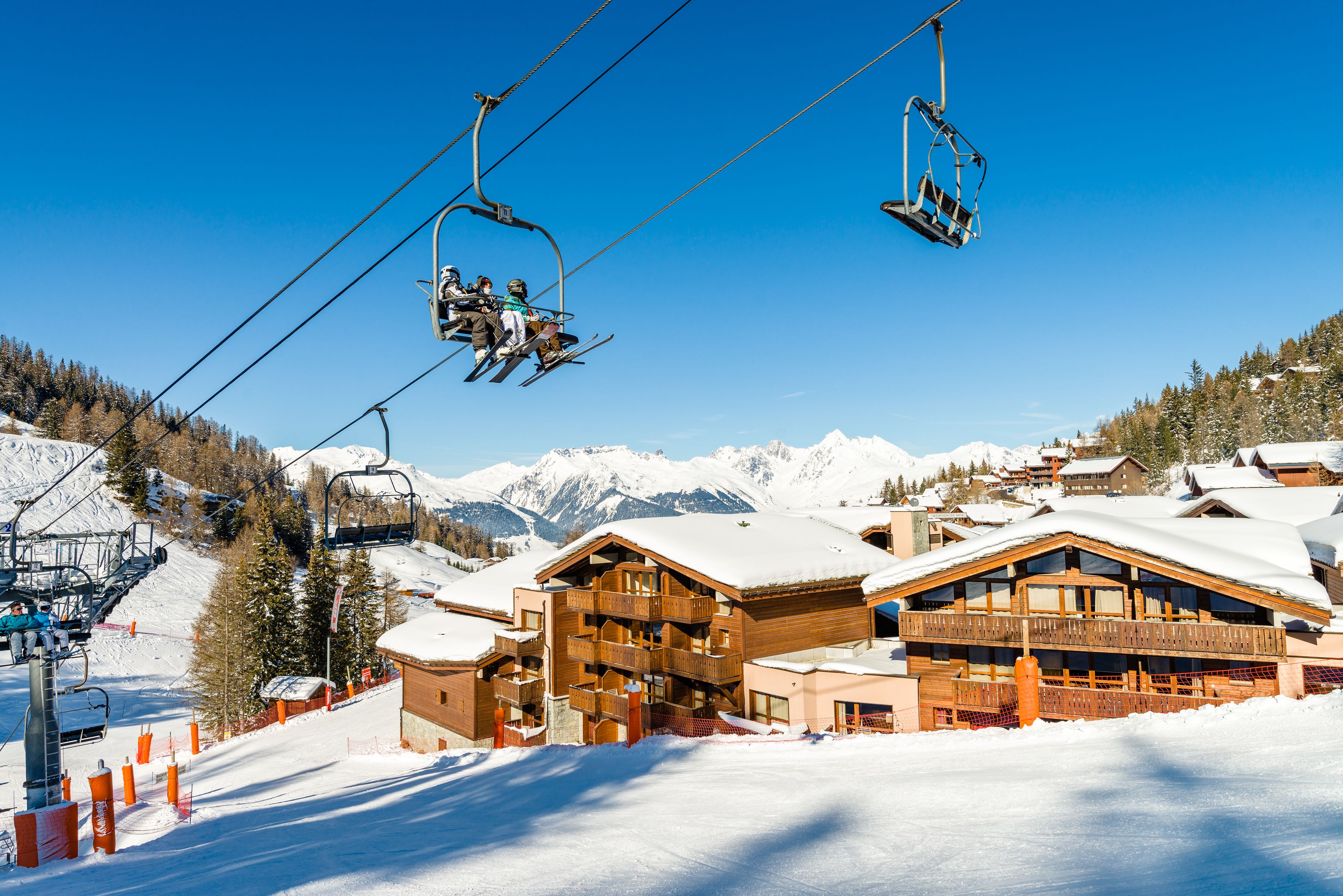 A ski lift with skiers, a wooden lodge with balconies, and snow-covered mountains in the background.