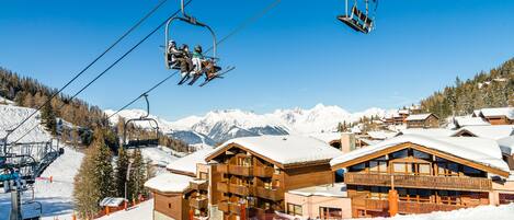 Eine Sesselbahn mit Skifahrern, eine Holzhütte mit Balkonen und schneebedeckte Berge im Hintergrund.
