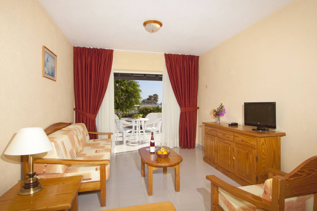 A room with a sofa, a table with a bottle and fruit, a TV on a wooden cabinet, and a view of the outdoors through a window with red curtains.