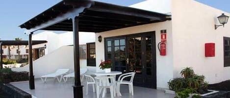 A covered outdoor seating area with white plastic chairs and tables, a black pergola, and a white building with a glass door and windows.