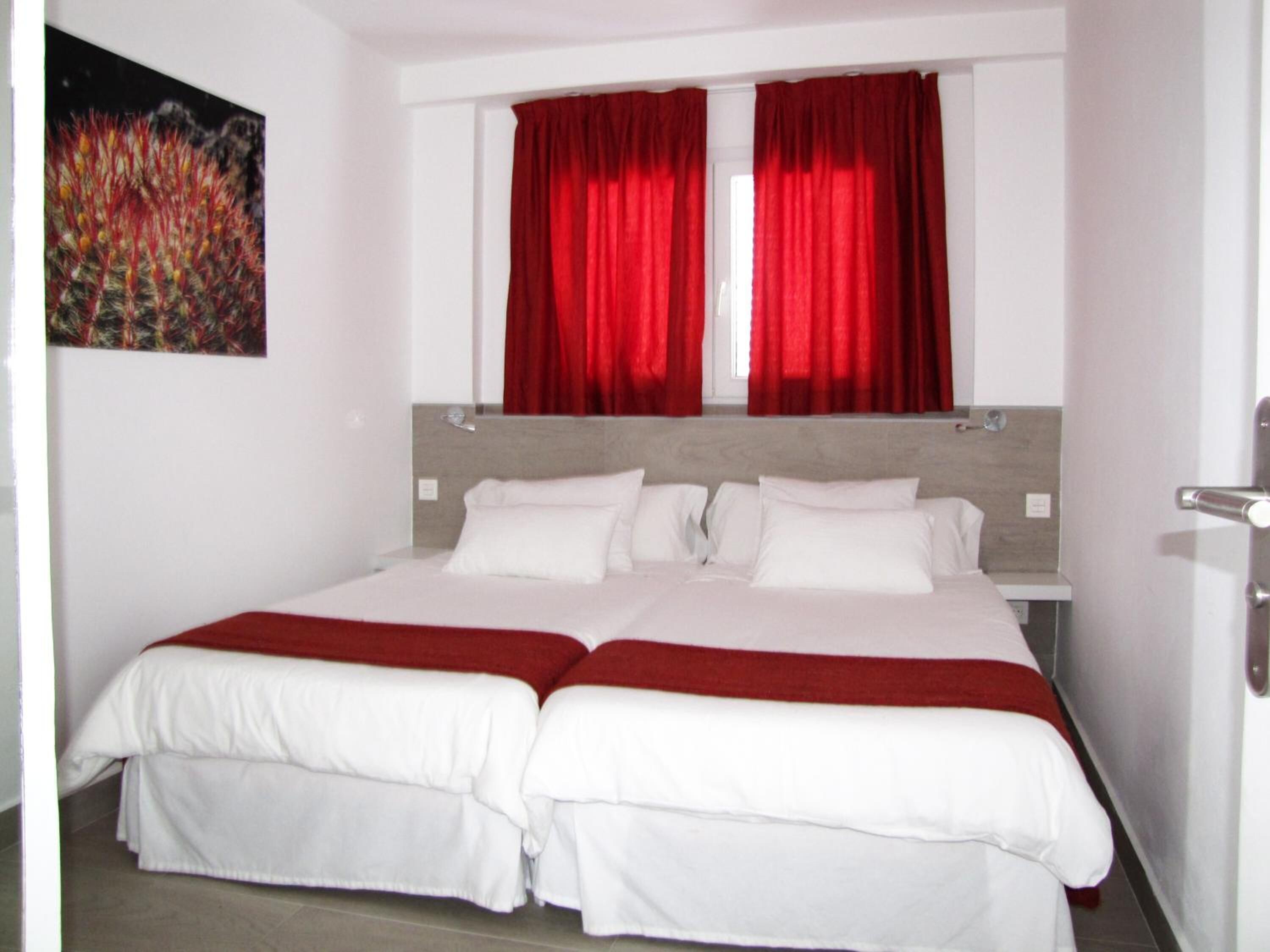 A double bed with white bedding and red accents, a window with red curtains, and a painting on the wall.