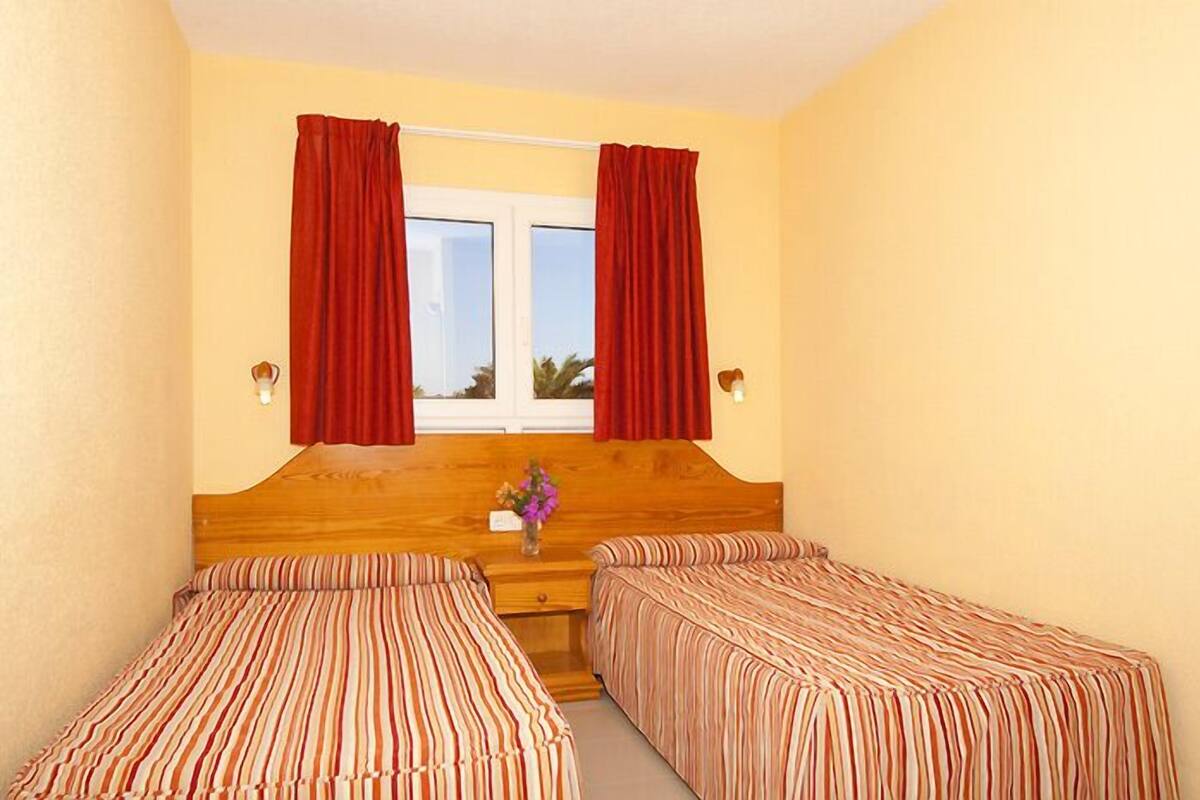 A room with two single beds, a wooden headboard, a window with red curtains, and a vase with flowers on the nightstand.
