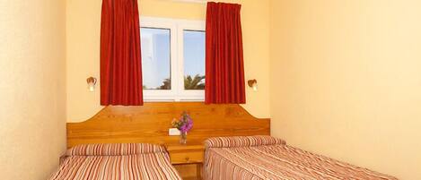 A room with two single beds, a wooden headboard, a window with red curtains, and a vase with flowers on the nightstand.