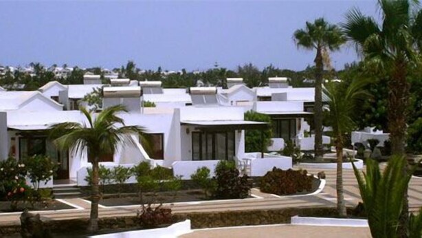 A row of modern white buildings with flat roofs, surrounded by palm trees and a clear sky.