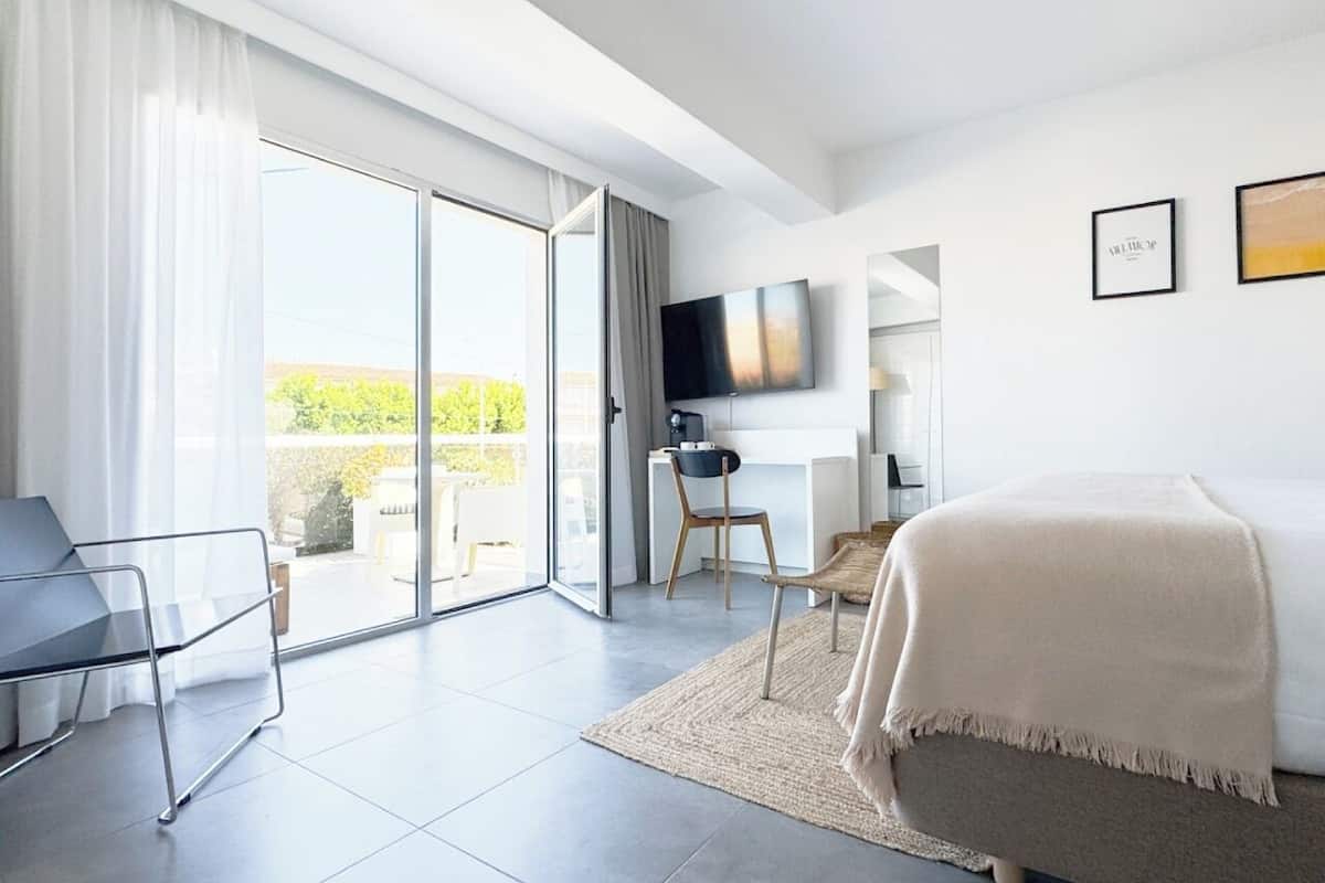 Quarto de hotel moderno com cama, escrivaninha, cadeira, televisão e porta de vidro deslizante para o exterior.