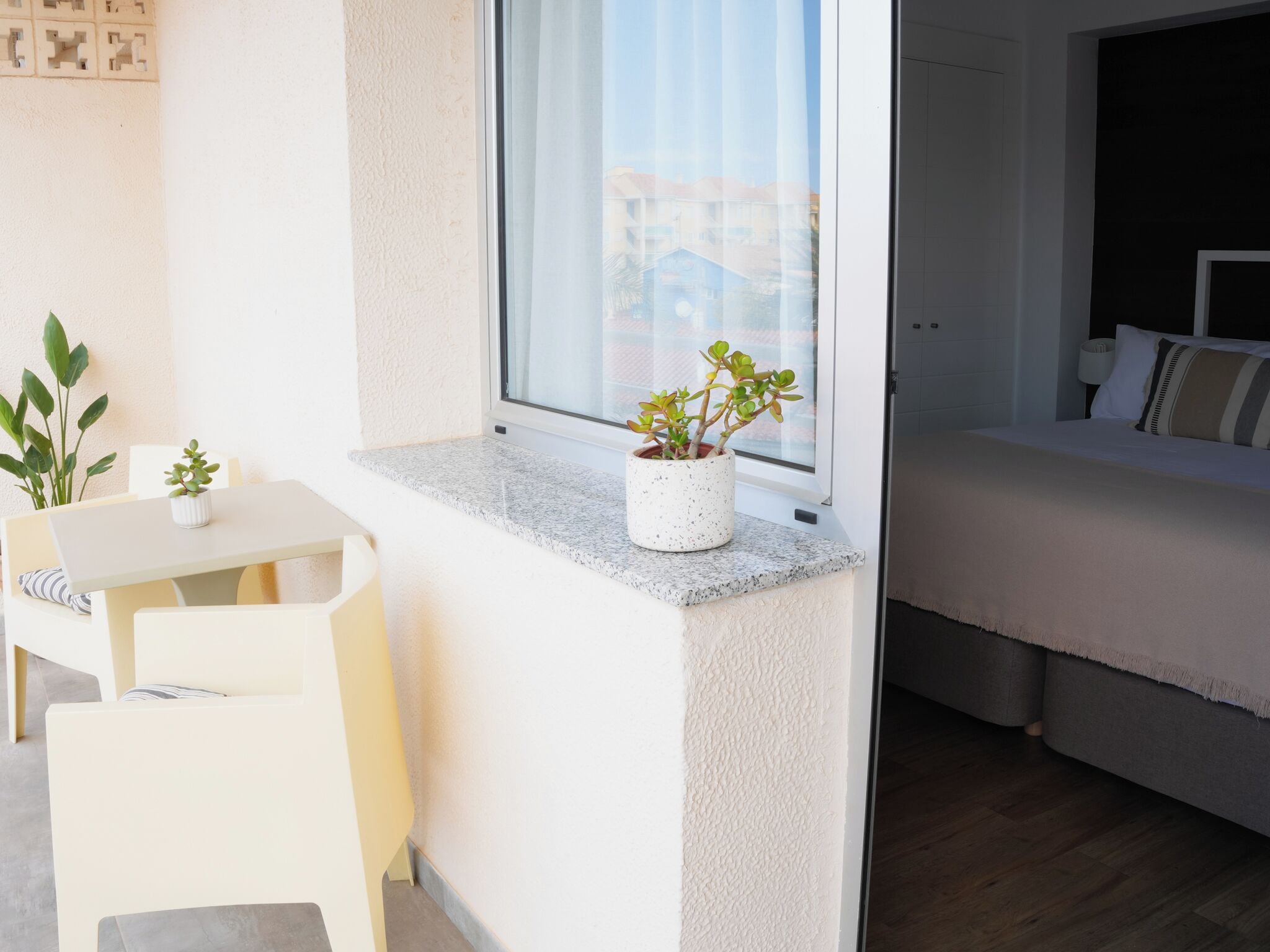 A small balcony with a table and chairs, a potted plant, and a view of buildings through a window.