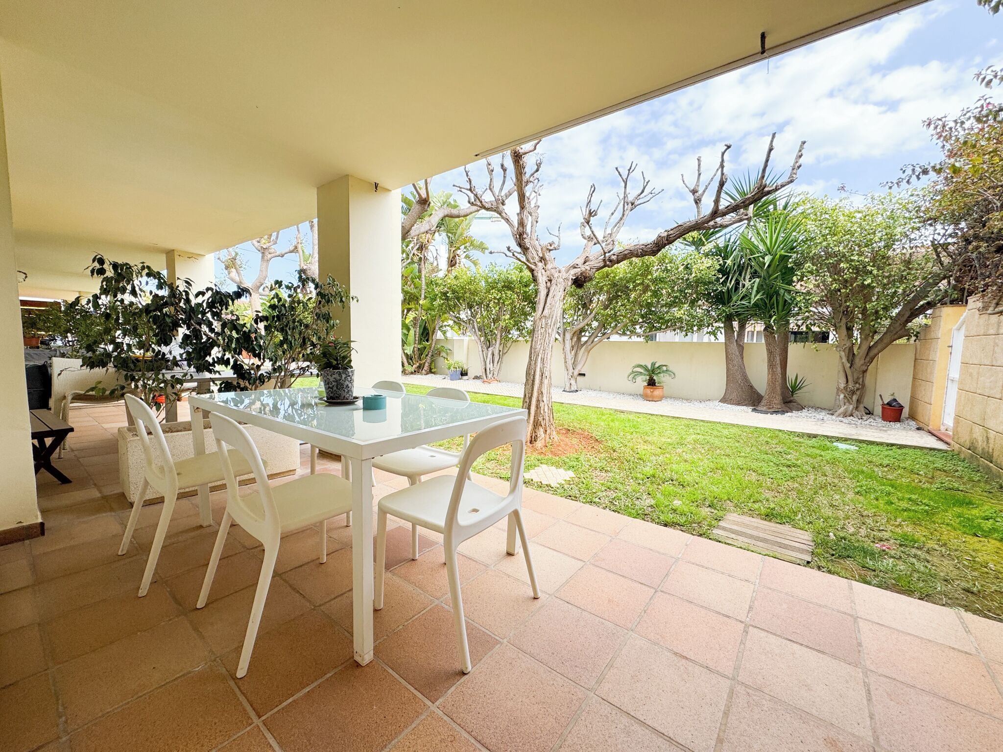 An outdoor patio with a glass table and white chairs, surrounded by a garden with various plants and trees.