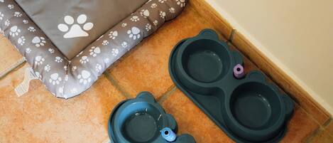 A pet bed with a paw print pattern and two pet bowls on a tiled floor.
