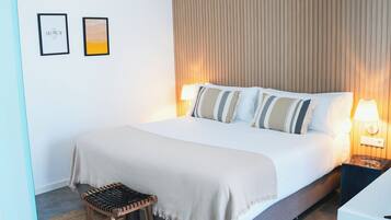 Una cama bien hecha con una colcha blanca y almohadas a rayas, un taburete de madera y dos lámparas de pared.