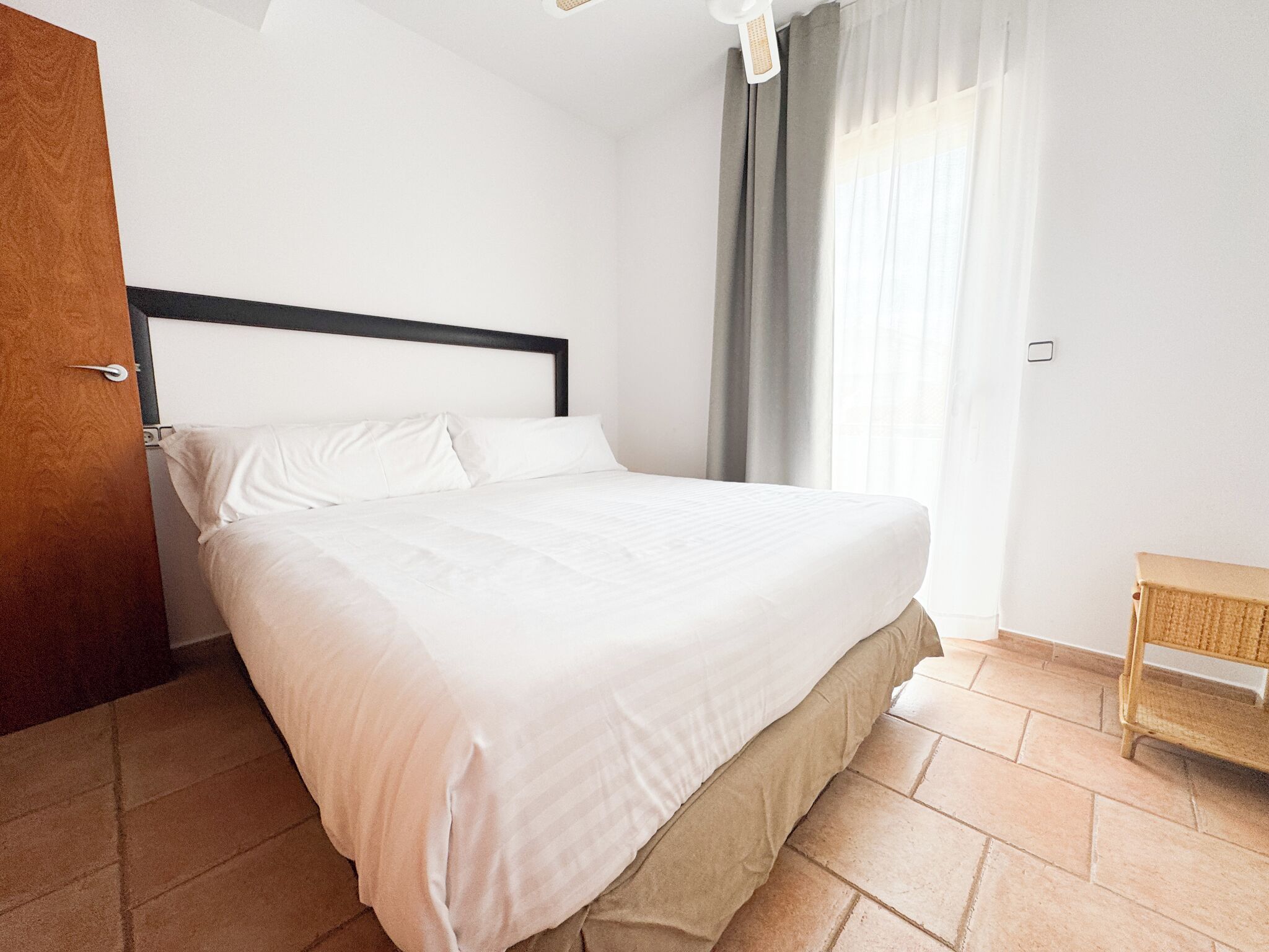 Una cama bien hecha en una habitación con una puerta de madera, un pequeño banco de madera y una ventana con cortinas.