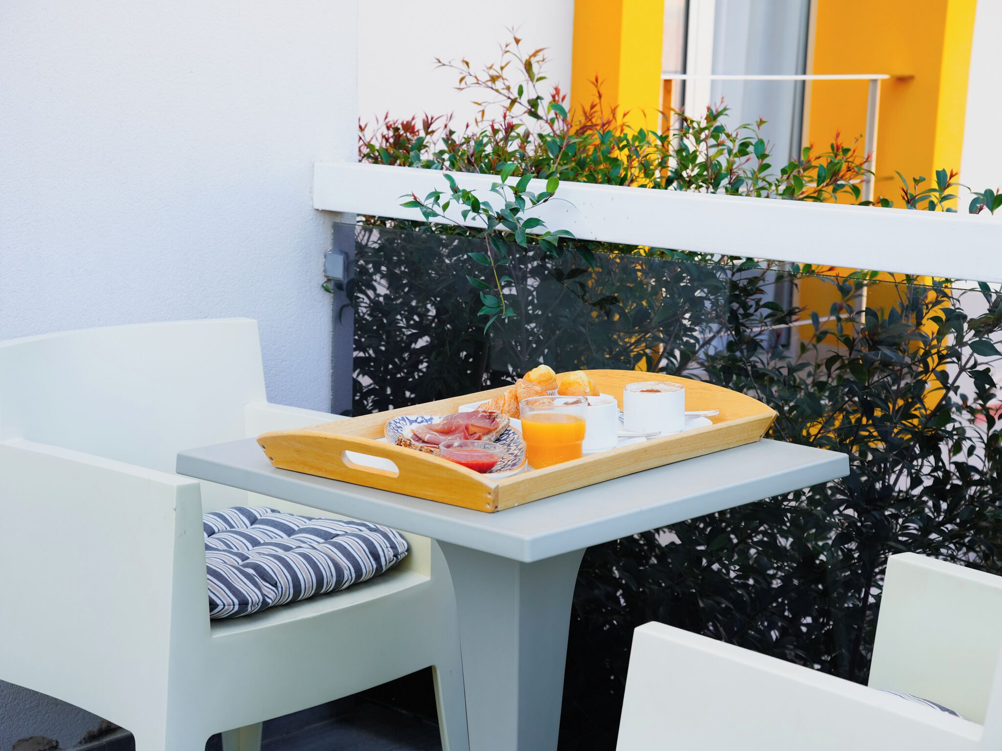 A breakfast tray with pastries, fruit, and beverages on a white table in a modern room with a white chair and a striped pillow.