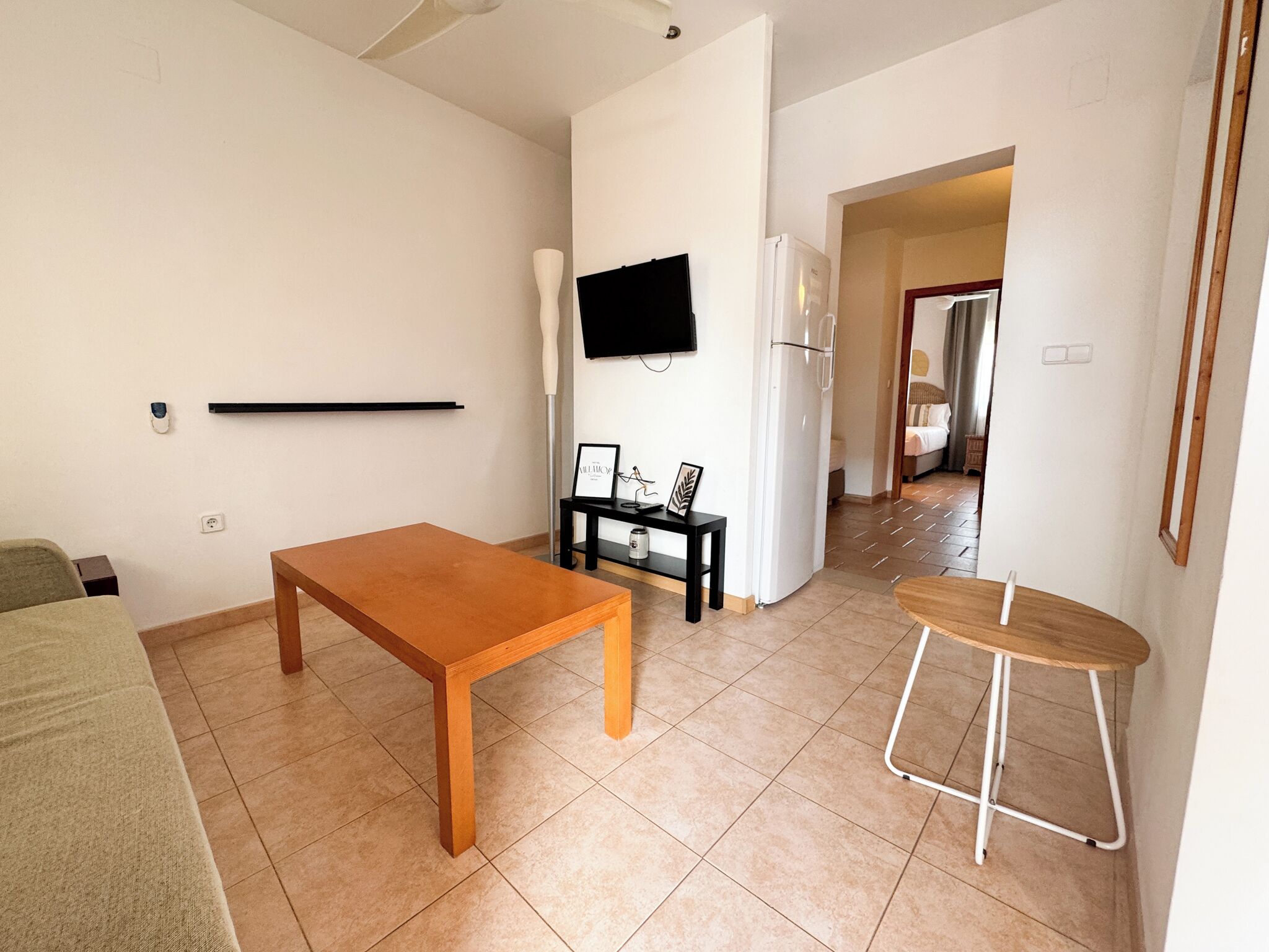 Una sala de estar con una mesa de madera, una mesita auxiliar negra con un marco de fotos, un refrigerador blanco y un televisor montado en la pared.