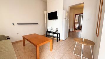 Una sala de estar con una mesa de madera, una mesita auxiliar negra con un marco de fotos, un refrigerador blanco y un televisor montado en la pared.
