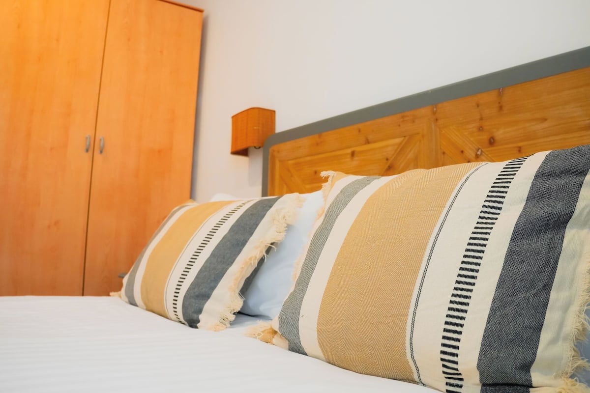 Quarto com guarda-roupa de madeira, cama com almofadas listradas e cabeceira de madeira.