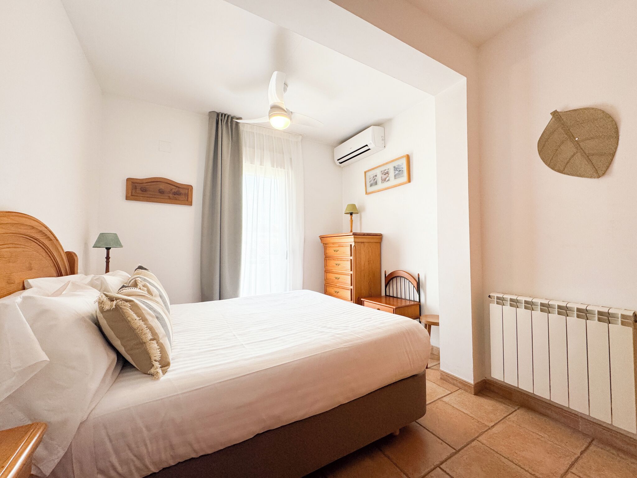 Een slaapkamer met een bed, een dressoir, een stoel, een lamp, een plafondventilator en een airconditioning.