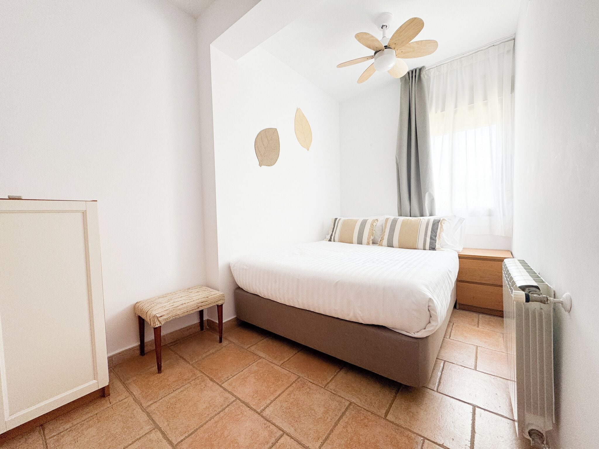 A bedroom with a bed, a small stool, a ceiling fan, a window with curtains, and a radiator.