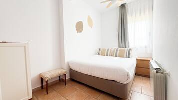 A bedroom with a bed, a small stool, a ceiling fan, a window with curtains, and a radiator.