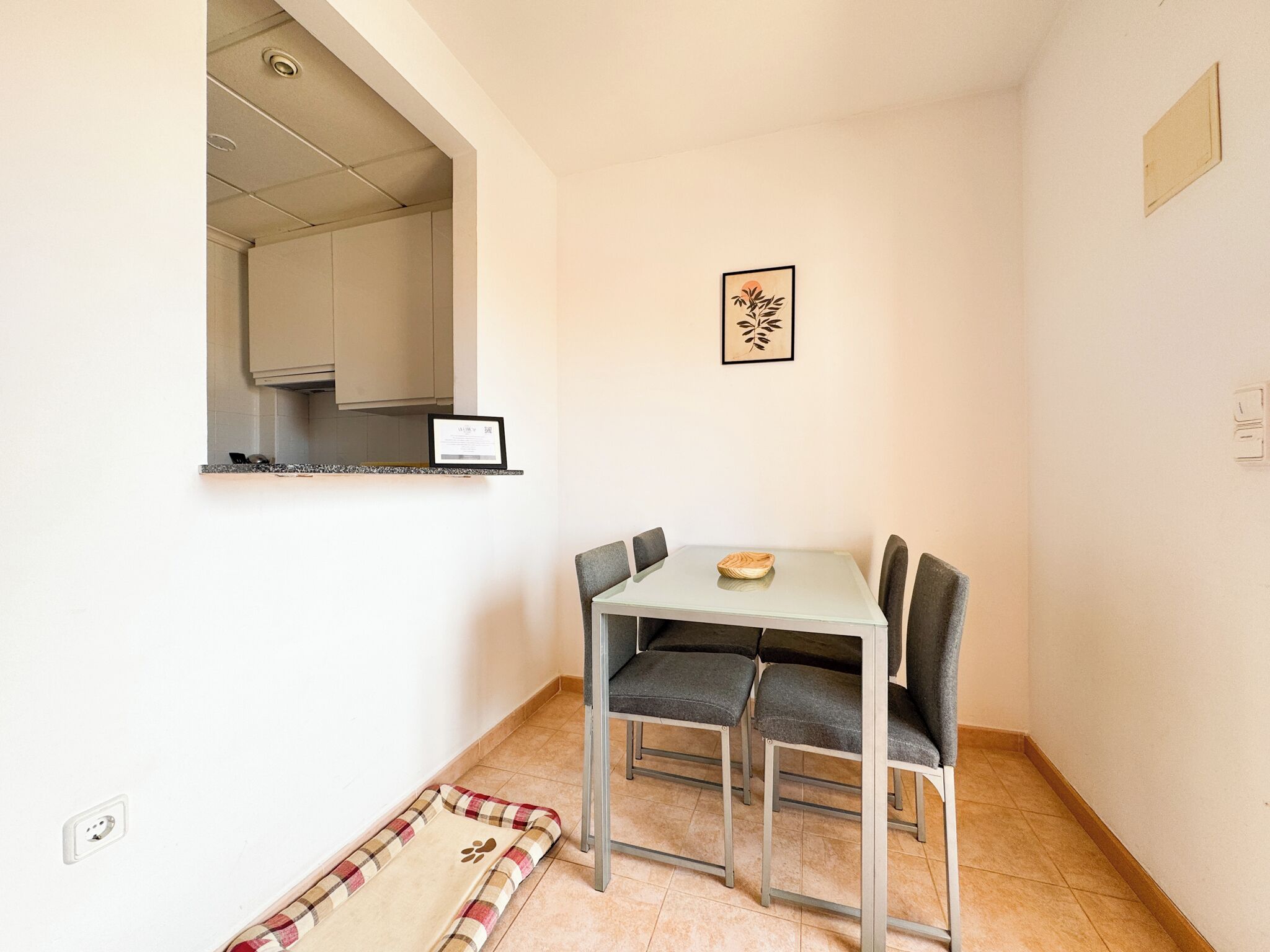 A small dining area with a table and four chairs, a framed picture on the wall, and a kitchen visible through an open doorway.
