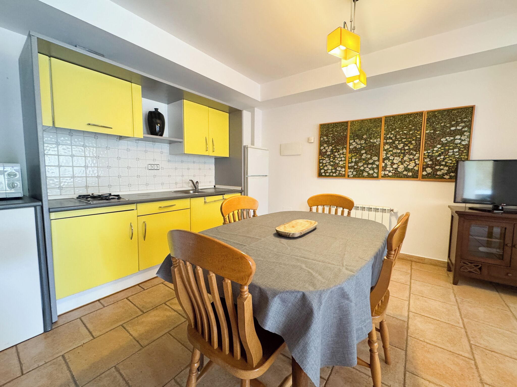 A compact kitchen with yellow cabinets, a dining table with chairs, a TV, and a floral artwork on the wall.