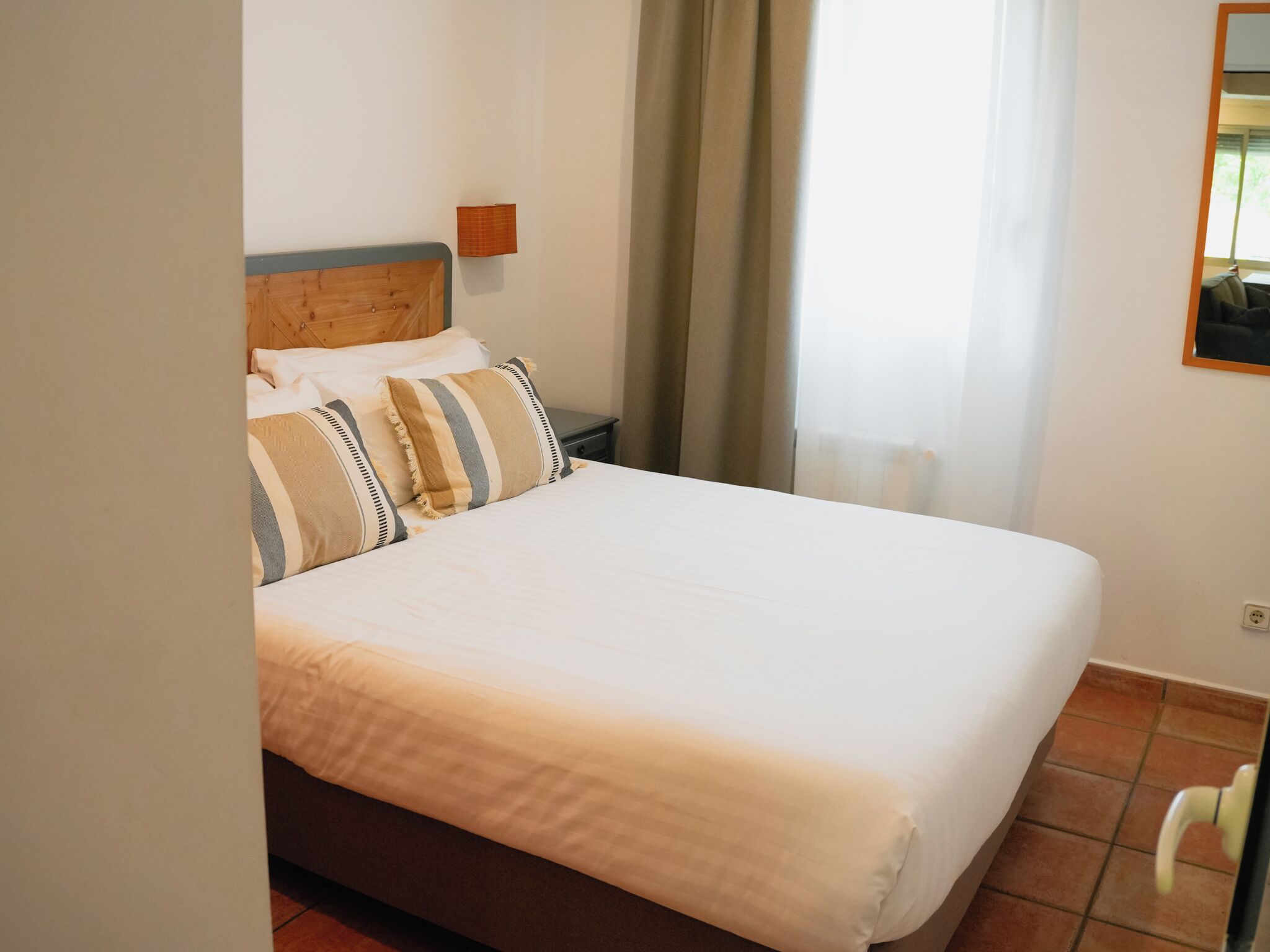A neatly made bed with striped pillows, a wooden headboard, a bedside table, and a window with curtains.