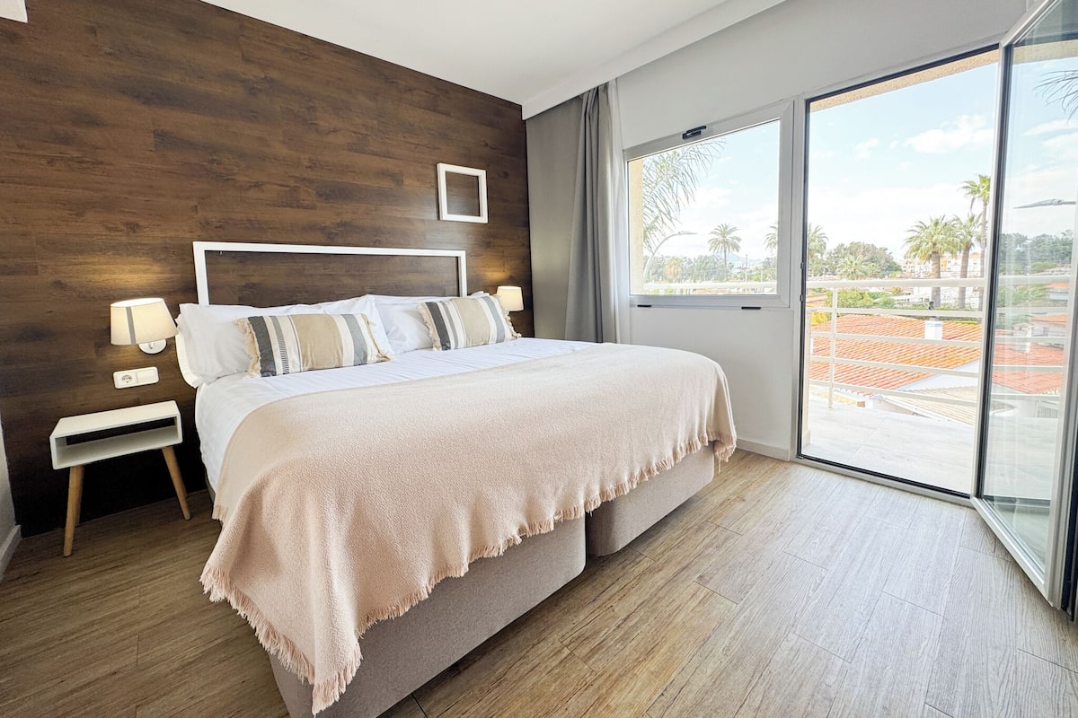 Quarto moderno com uma cama grande, painéis de parede de madeira, uma mesinha de cabeceira, abajur e uma grande janela com vista para palmeiras e edifícios.