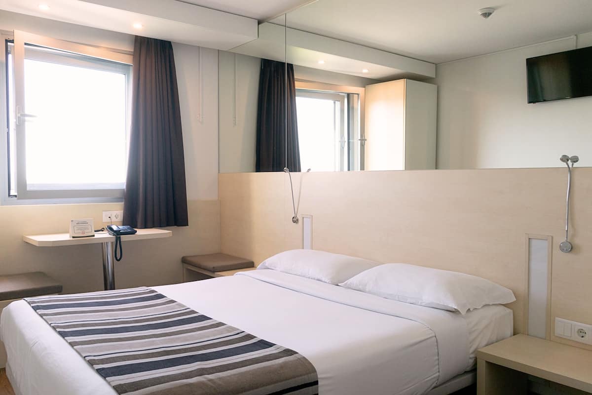A hotel room with a bed, a bedside table, a mirror, a TV mounted on the wall, a window with curtains, and a shower area.