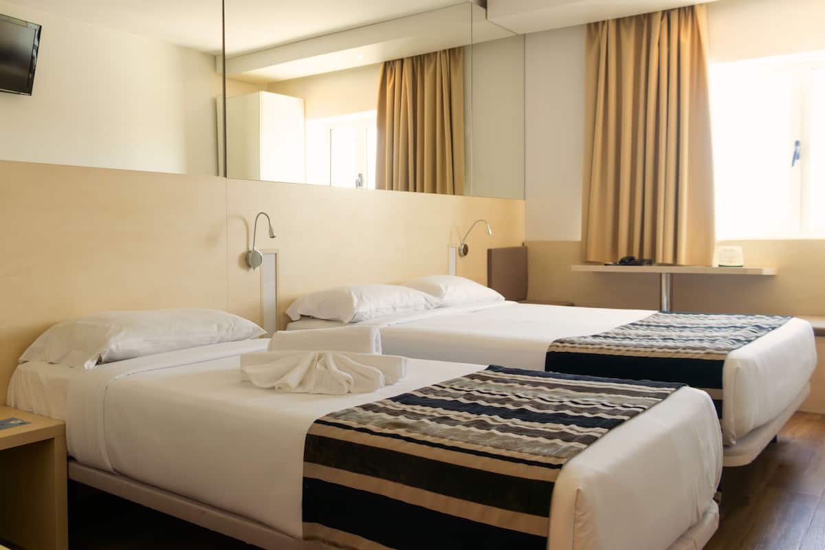 A hotel room with two beds, a bedside table, a mirror, a window with curtains, and a flat-screen TV.