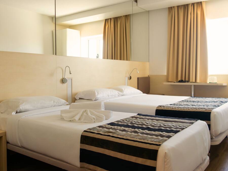 A hotel room with two beds, a bedside table, a mirror, a window with curtains, and a flat-screen TV.