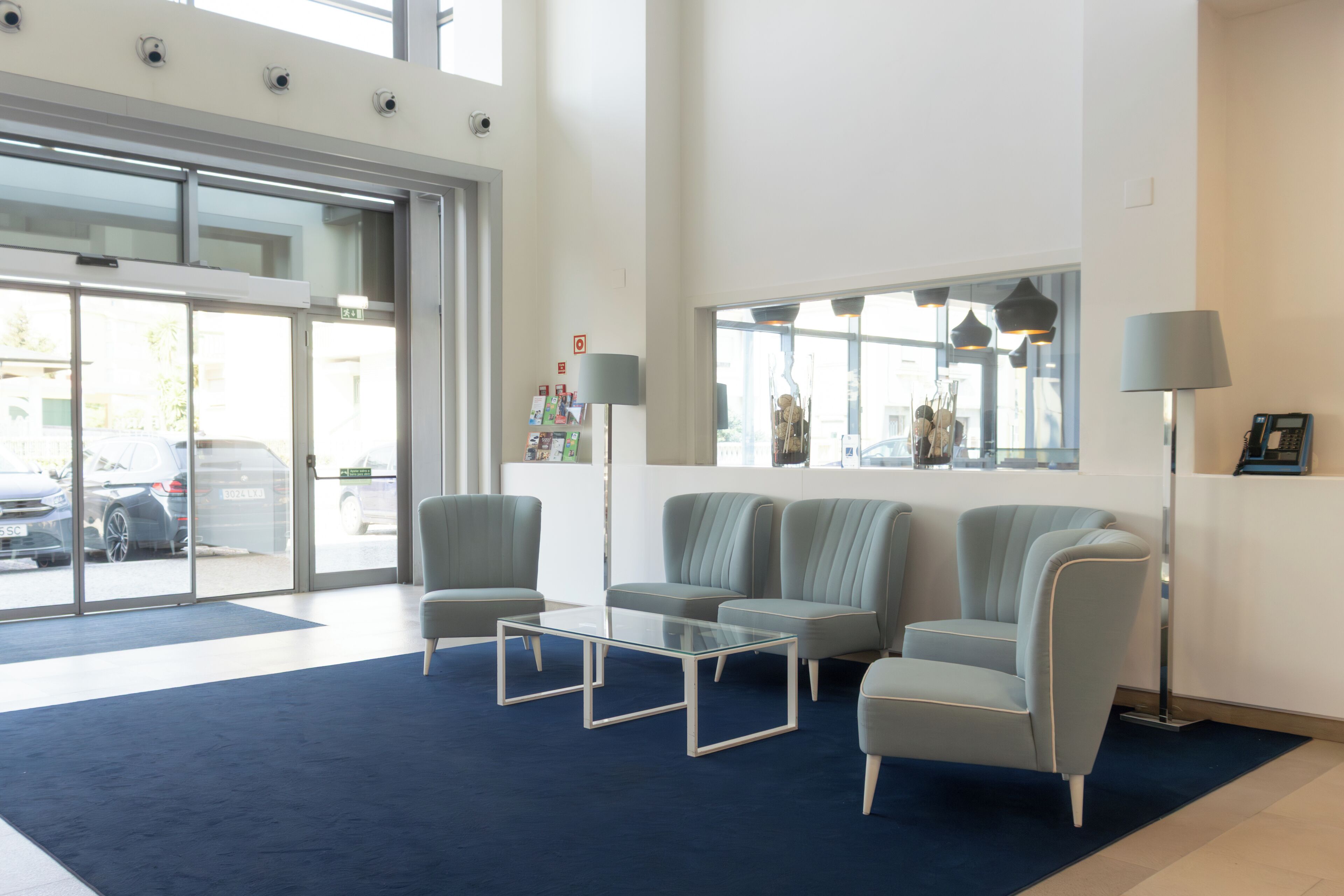 Un moderno vestíbulo con entrada por puerta de vidrio, alfombra azul y una zona de estar con sofá y sillones.