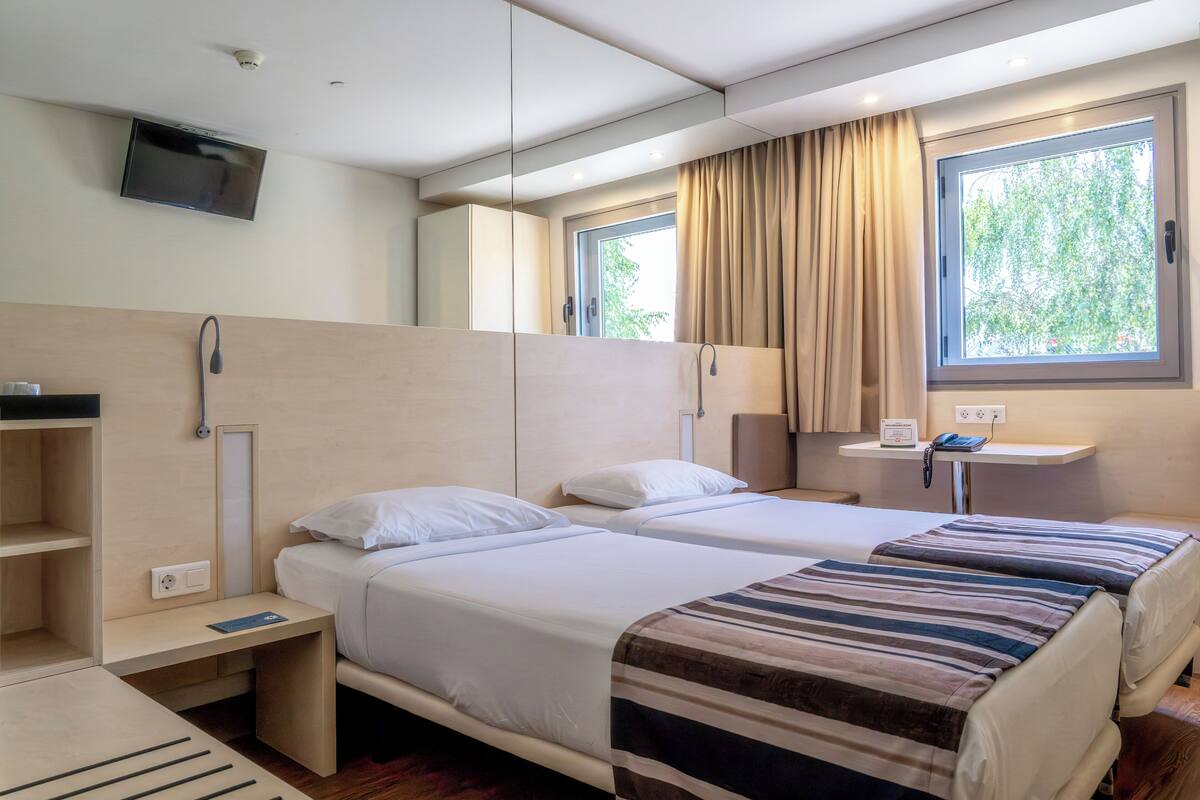 A hotel room with two beds, a TV, a window with curtains, a bedside table, and a mirror.