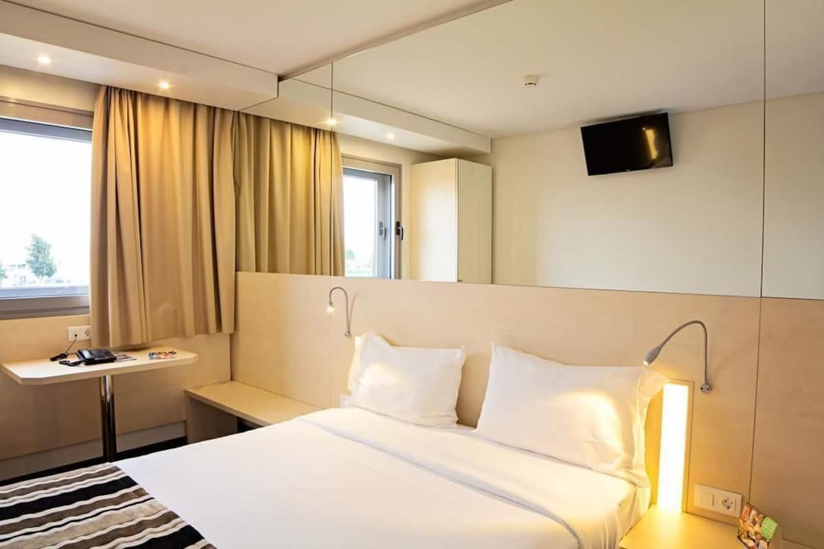 A hotel room with a bed, bedside lamp, a small table with a phone, a TV mounted on the wall, and a window with curtains.