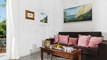 Una sala de estar con un sofá negro, cojines rosa, una mesita de centro con fruteros y un cuadro en la pared.