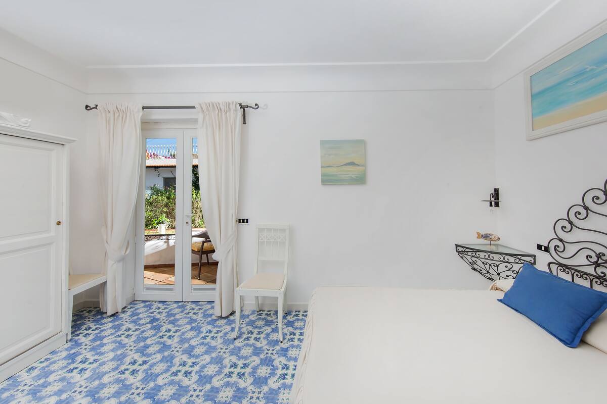 Une chambre avec un lit, une chaise, une petite table et une vue sur un espace extérieur, accessible par une porte ouverte.