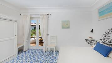 A bedroom with a bed, a chair, a small table, and a view through an open door to an outdoor area.