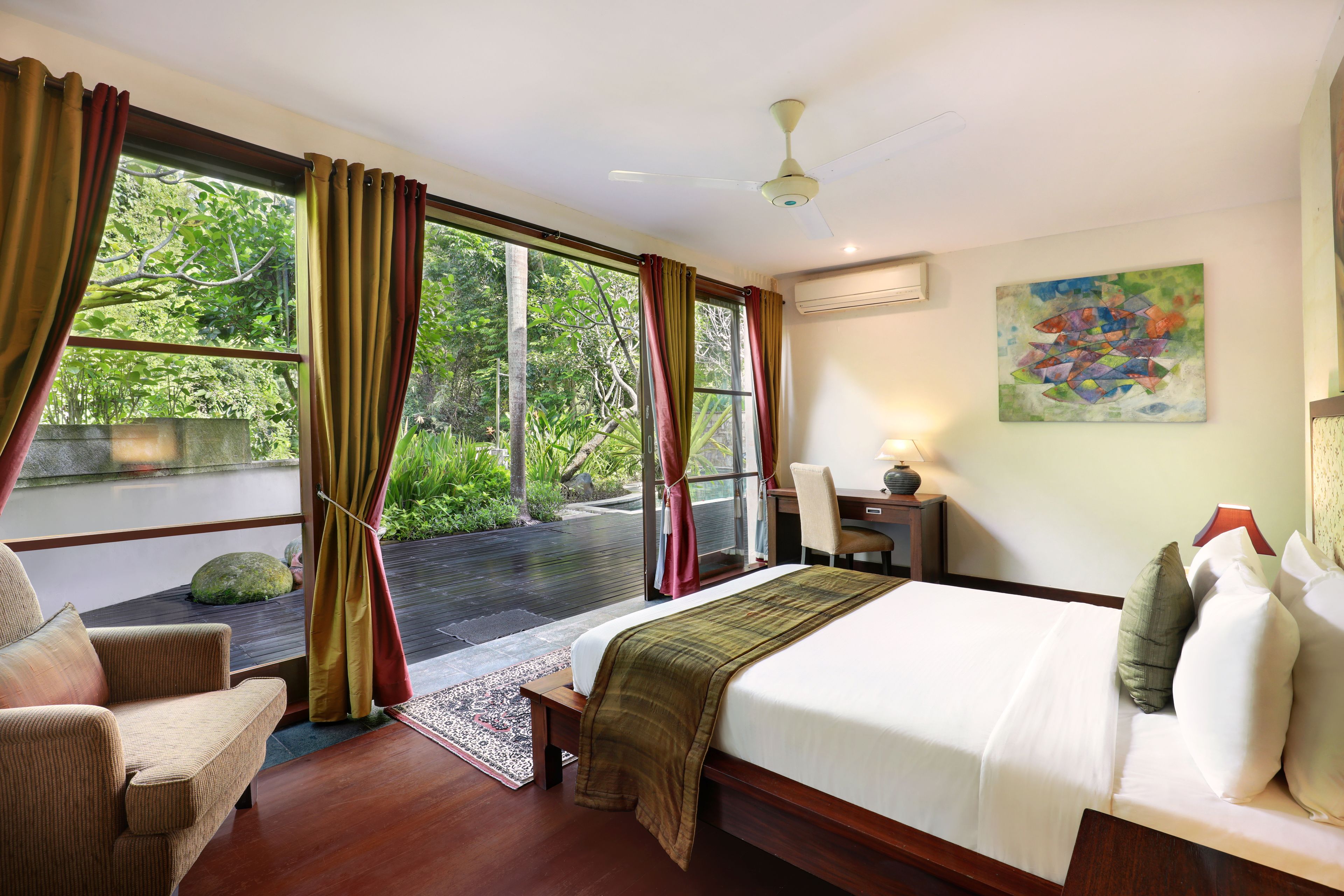 Une chambre d’hôtel avec un grand lit, une chaise, un bureau et une vue sur une végétation luxuriante par les fenêtres.