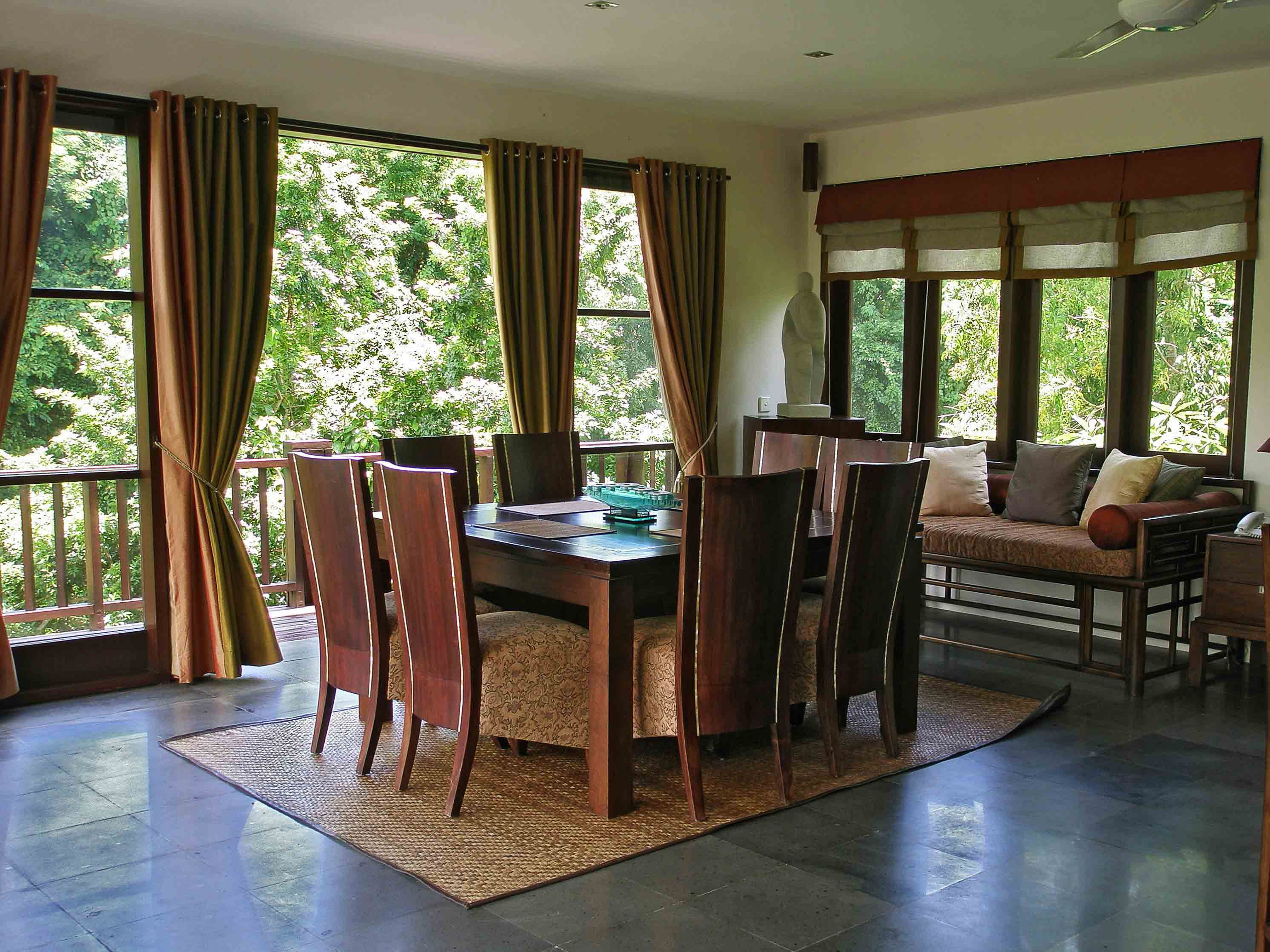 A dining area with a wooden table and chairs, a sofa, and large windows with curtains.