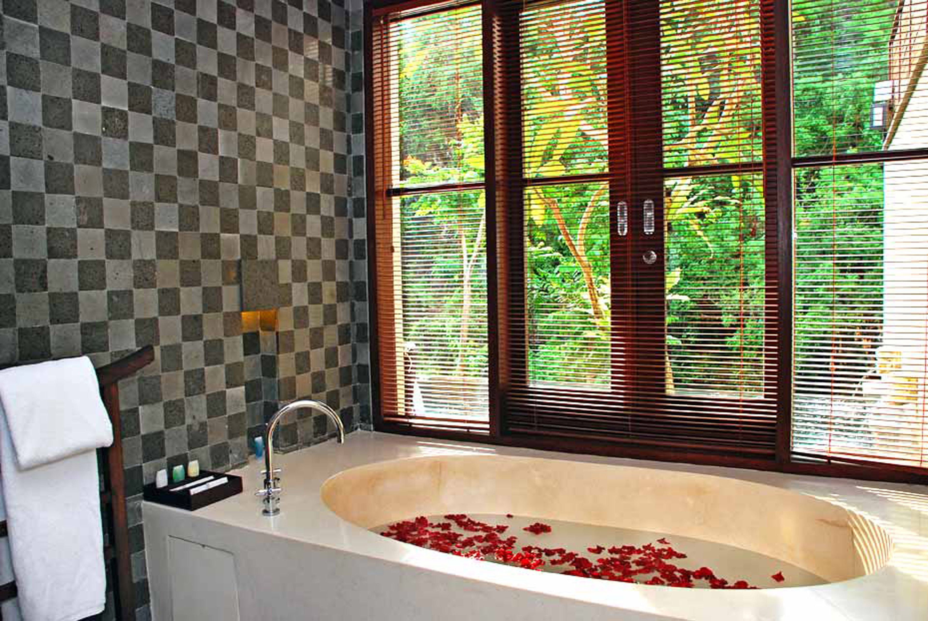 A bathroom with a checkered wall, a bathtub with rose petals, and a window with blinds.