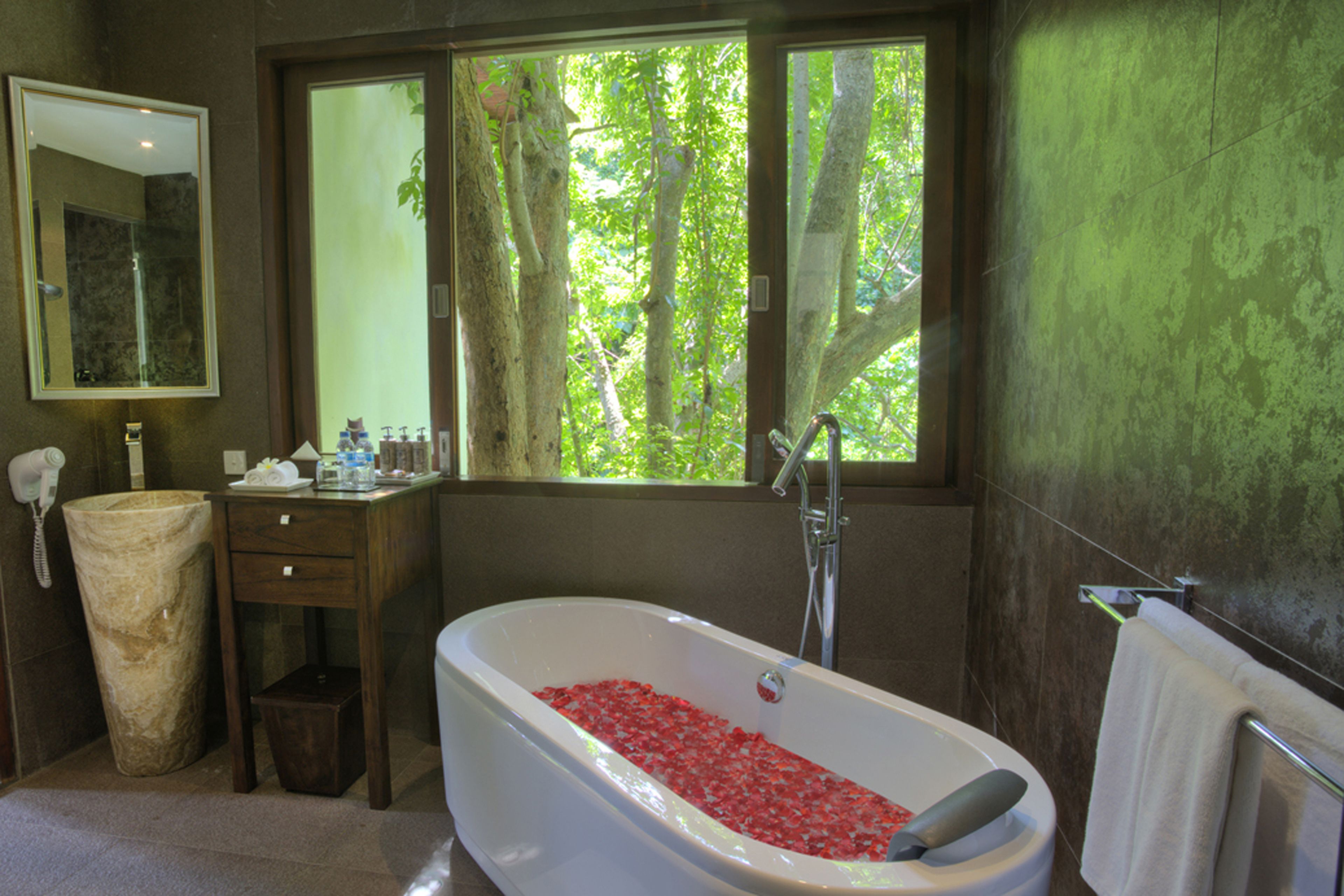 A bathroom with a bathtub filled with red flowers, a sink, a mirror, and a window with a view of trees.