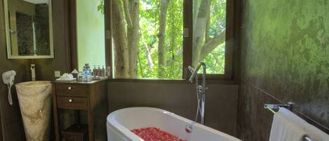 A bathroom with a bathtub filled with red flowers, a sink, a mirror, and a window with a view of trees.