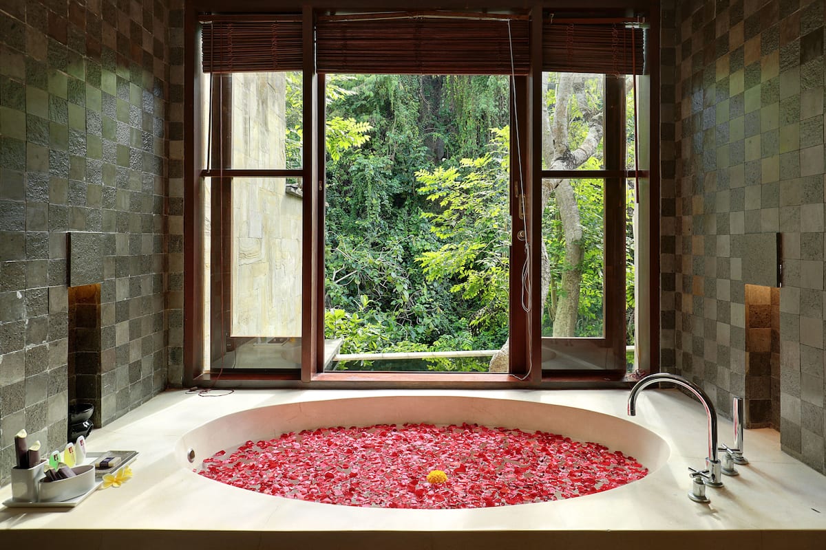 Une salle de bain dotée d’une grande fenêtre, d’une baignoire remplie de pétales de roses rouges et d’un mur en pierre.