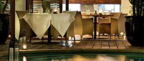 Zona junto a la piscina con sillas y mesas de mimbre, piscina y plantas en macetas.