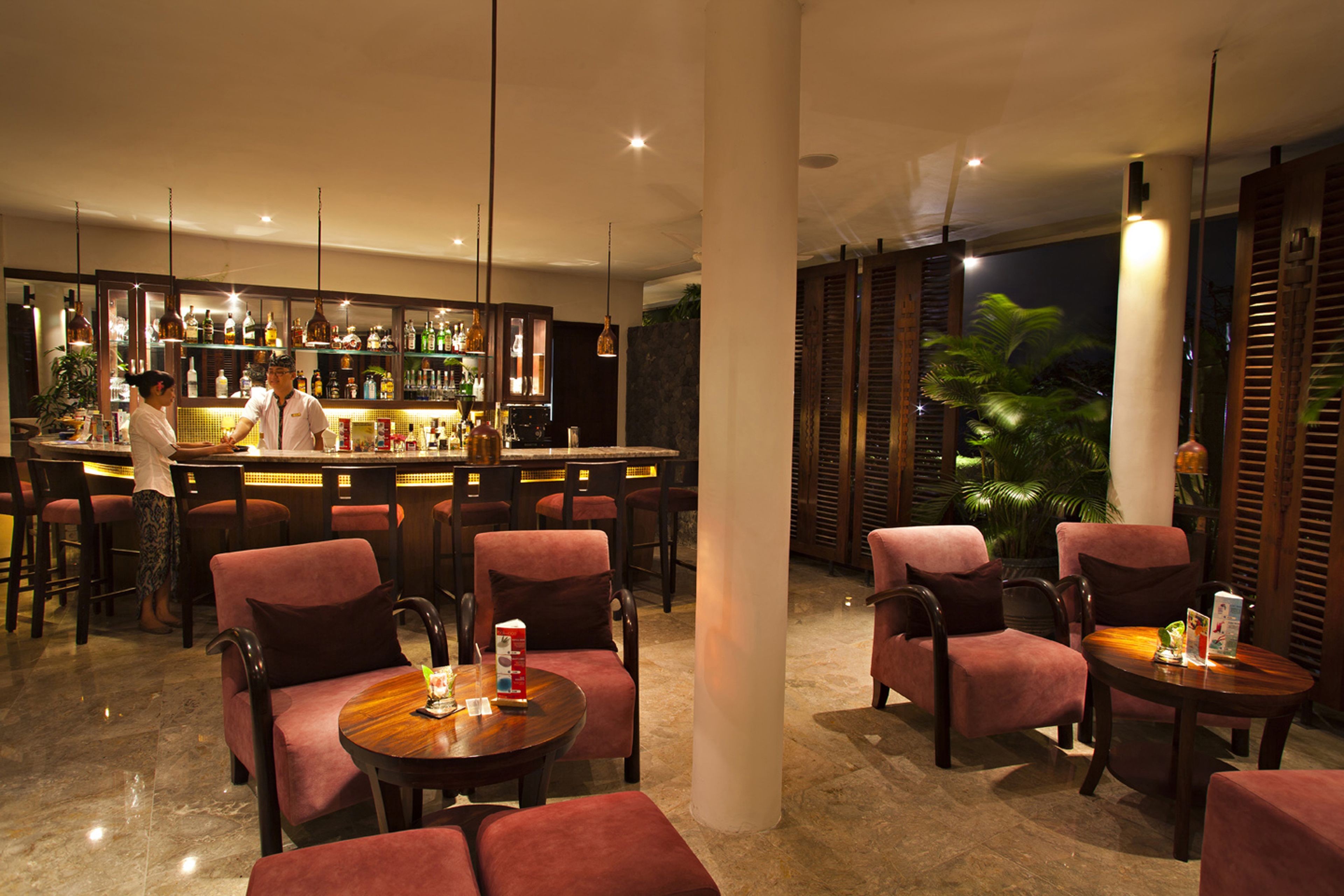 Un espace bar avec un barman, des chaises à l’assise rembourrée de couleur rouge et un comptoir où sont exposées différentes bouteilles.