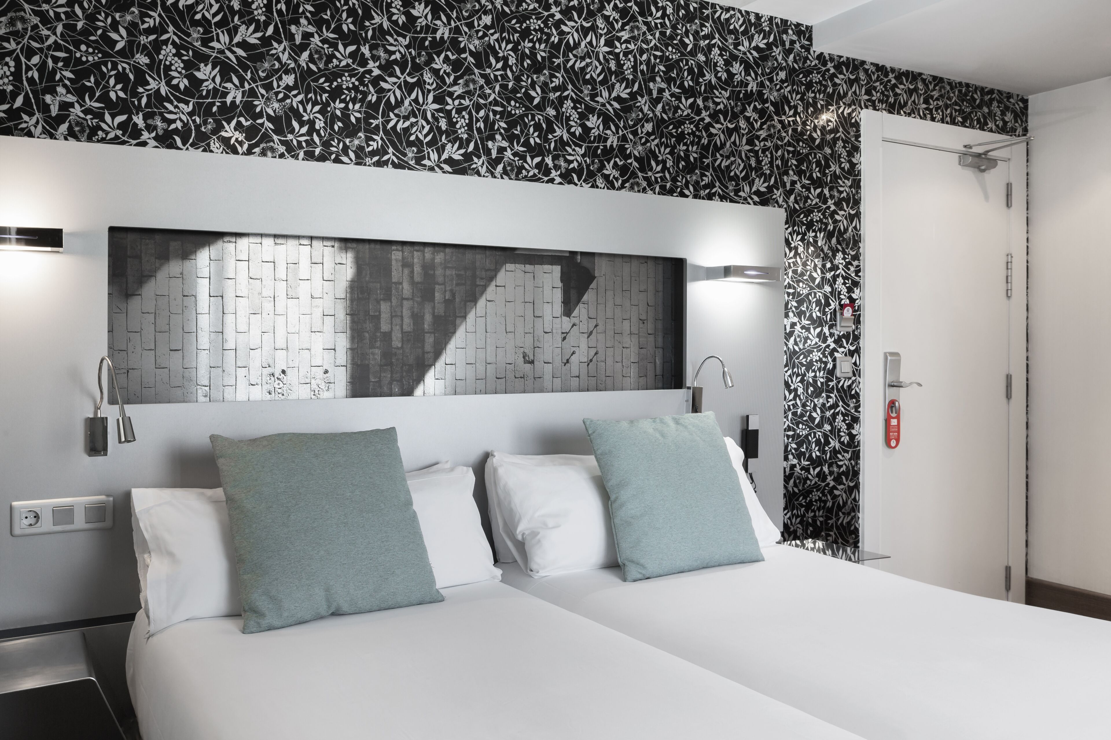 A hotel room with a bed, two pillows, a nightstand, a wall-mounted light, a mirror, and a door.