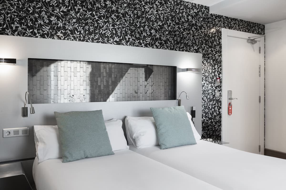 Hotellihuone, jossa on sänky, kaksi tyynyä, yöpöytä, seinään kiinnitetty valaisin, peili ja ovi.