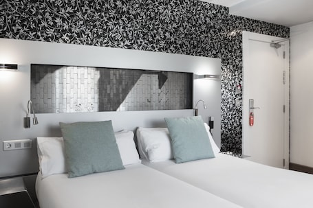 A hotel room with a bed, two pillows, a nightstand, a wall-mounted light, a mirror, and a door.