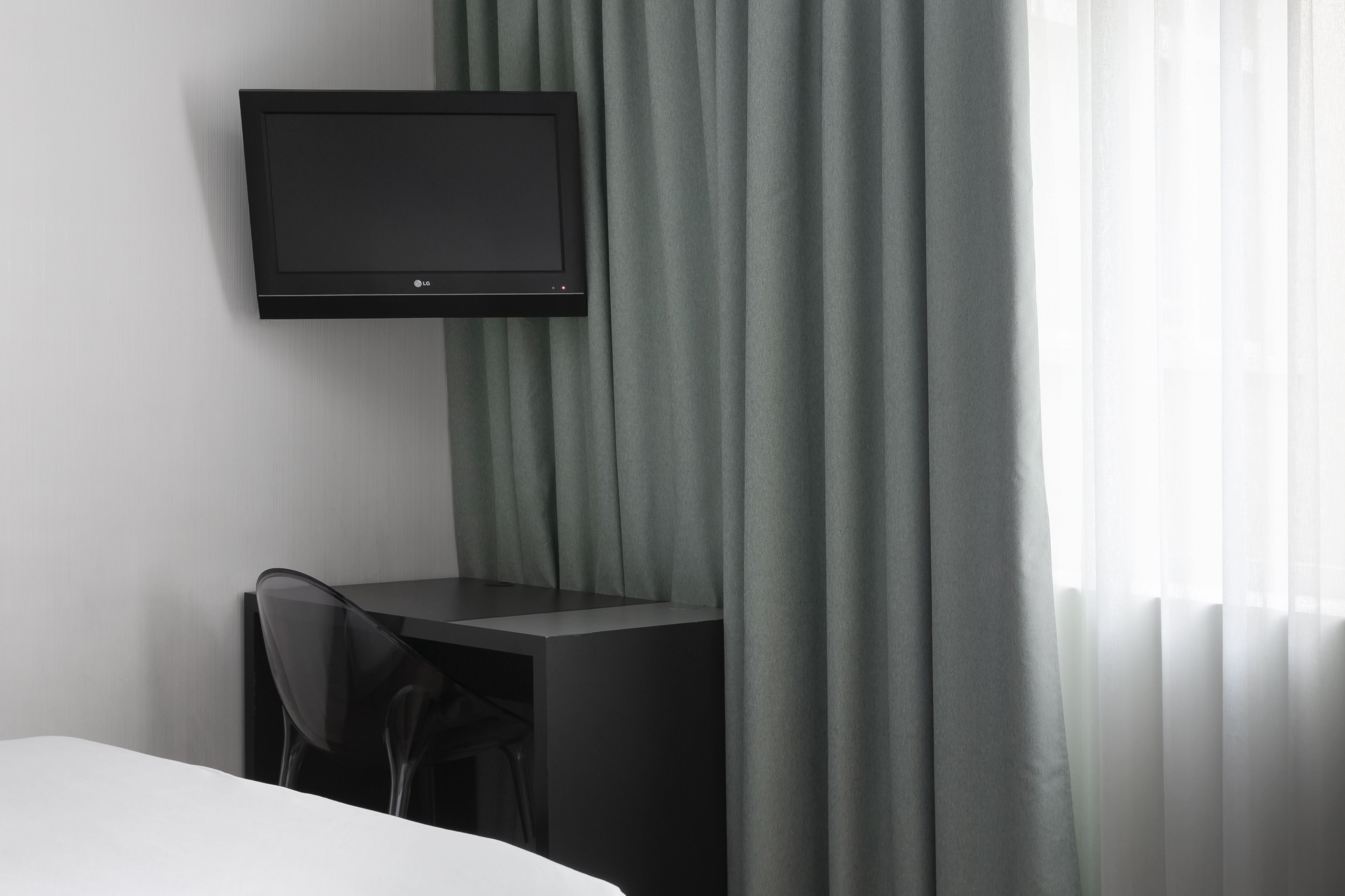 A modern hotel room with a flat-screen TV mounted on the wall, a small desk with a chair, and a large window with sheer curtains.