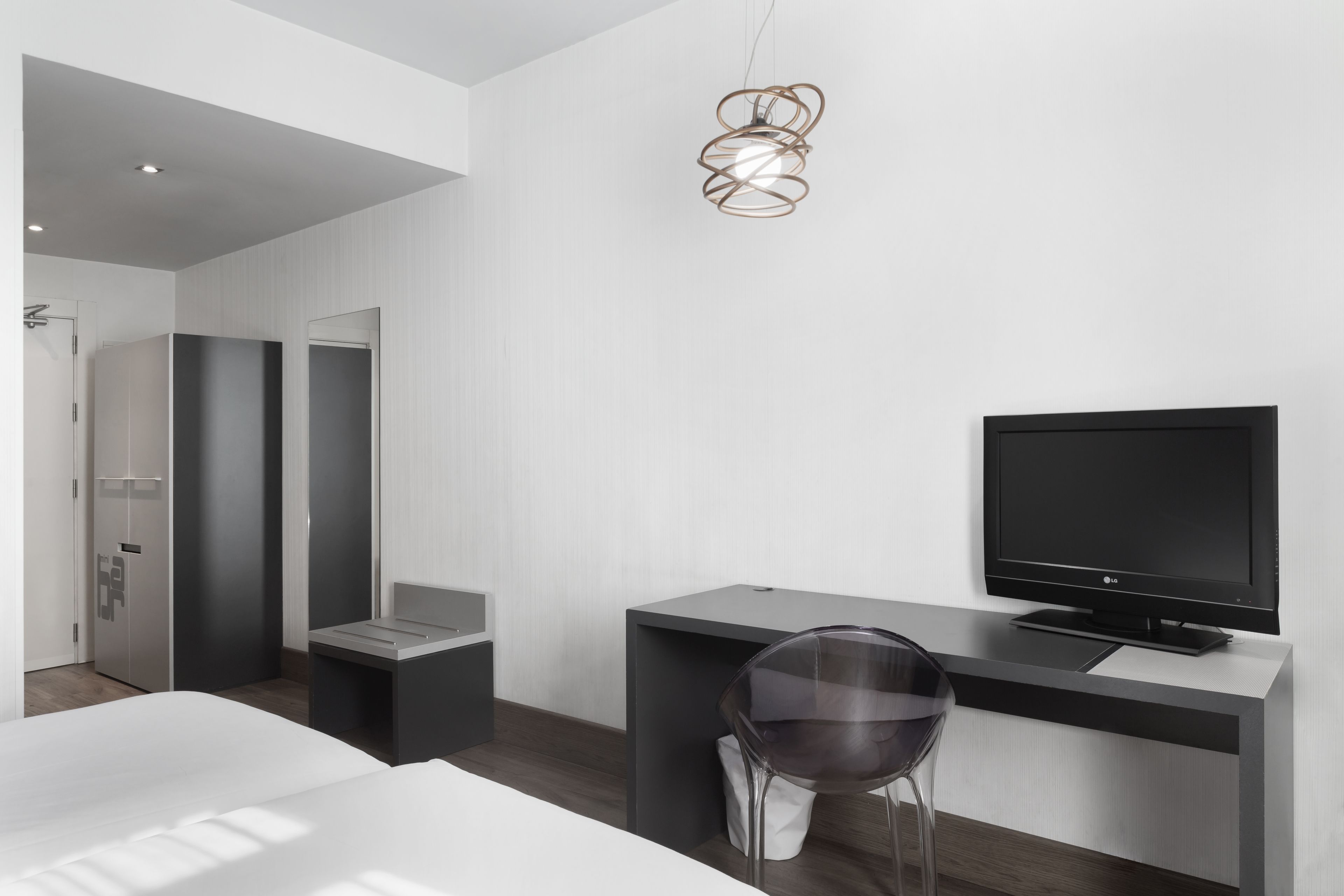 A modern hotel room with a bed, a desk with a TV, a chair, and a unique pendant light.